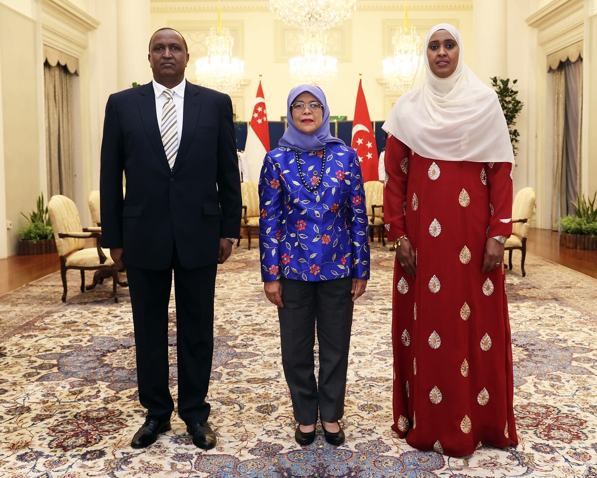 Three people stand in formal attire with Singapore flags behind them.